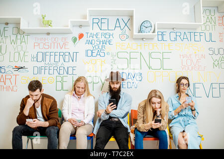 Group of diverse people using smart phones while sitting together in a row on the wall background indoors Stock Photo