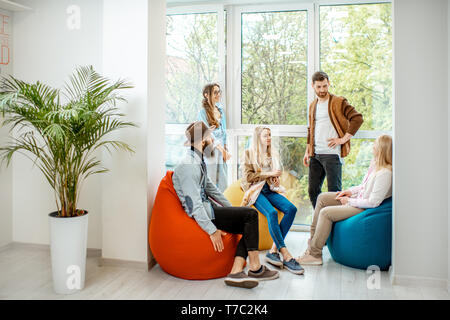Group of diverse people having a conversation during the coffee break sitting on the colorful poufs near the window in the office Stock Photo