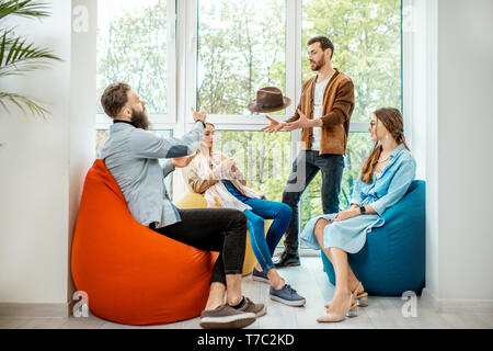 Group of diverse people having a conversation during the coffee break sitting on the colorful poufs near the window in the office Stock Photo