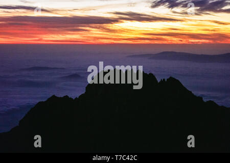 Orange sun is rising on the horizon, light falling on the clouds near the mountain peaks. Black sharp rocks. Adventures and trip in Africa. Stock Photo