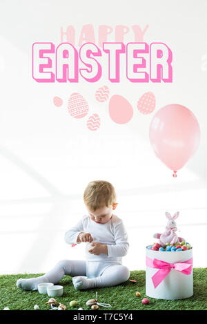 cute baby sitting near box with colorful quail eggs, toy rabbit and air balloon on white background with happy Easter lettering Stock Photo