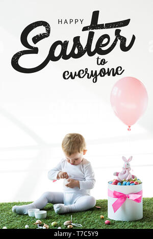 cute baby sitting near box with colorful quail eggs, toy rabbit and air balloon on white background with happy Easter to everyone lettering Stock Photo