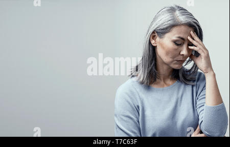 Serious Middle-Aged Woman Touches Her Forehead and Thinking About Something. Upseted Asian Woman With Grey Hair Stands And Touches Her Forehead Thinki Stock Photo