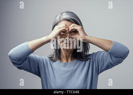 Beautiful Grey-Haired Woman with Dark Skin Squints Her Eyes While Looking Through Her Binoculars Like Hands. Attractive Asian in Light-Blue Sweater Po Stock Photo