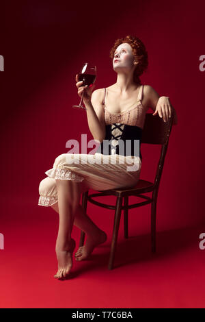Medieval redhead young woman as a duchess in black corset and night clothes sitting on the chair on red background. Drinking red wine. Concept of comparison of eras, modernity and renaissance. Stock Photo