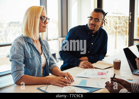 Two diverse young business colleagues smiling and discussing paperwork together while sitting at a desk in an office Stock Photo
