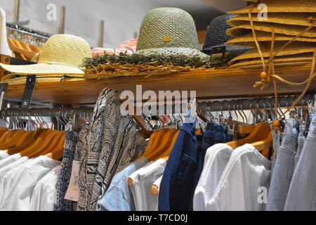 Dress rack full of wooden hangers with shirts and trousers and straw summer hats on shelf above. Stock Photo