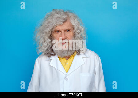 Old long-haired man wearing doctor's gown studio isolated on blue wall looking camera thoughtful Stock Photo