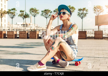 Young alternative girl skater wearing cap and sunglasses sitting on penny board on the city street looking up cool Stock Photo