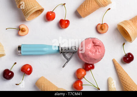 Homemade cherry ice cream scoop in spoon with waffle cones and ripe fresh cherries on white table flatlay top view