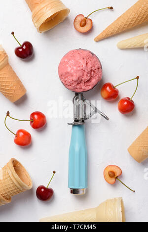 Cherry ice cream scoop in spoon with waffle cones and ripe fresh cherries on white table flatlay