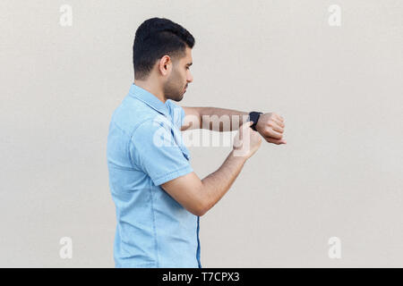 Profile side view portrait of attentive handsome young bearded man in blue shirt standing and using his smart watch, touching at screen. indoor studio Stock Photo