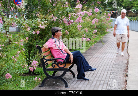 Man sitting on bench to relax while another male with white hair walking for do exercise in park, healthy lifestyle of people at big city, Vietnam Stock Photo