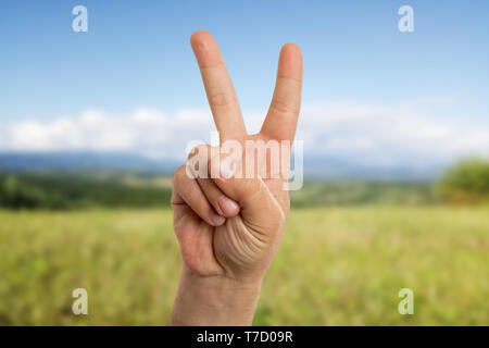 Pacifist man showing peace or victory sign using index and middle fingers with green field as background Stock Photo