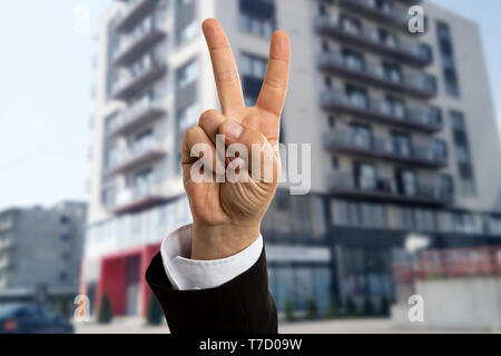 Male real estate agent showing victory or peace sign in front of apartment building for rent closeup Stock Photo