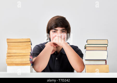 Exhausted stressed and angry teenage school boy sitting at the table between pile of books with his hands on his cheeks Stock Photo