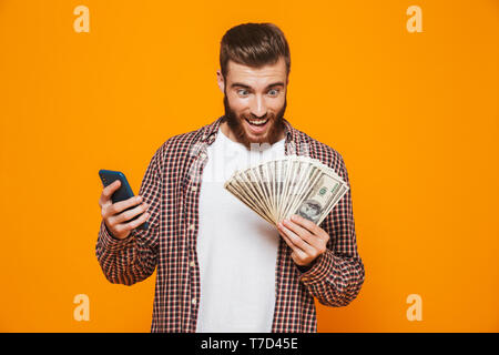 Portrait of a cheerful young man wearing casual clothes standing isolated over yellow background, holding mobile phone, showing money banknotes Stock Photo