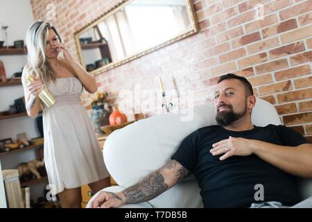 Man with tattoo sitting in armchair and woman having golden bottle of champagne in background. Relationship between husband and wife at home. Stock Photo