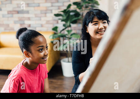 Young teacher giving African American kid an art lesson and teaching how to paint on easel inside living room in natural ambient light Stock Photo