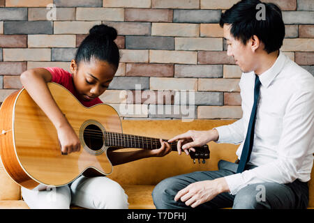 Young Asian male teacher giving a guitar lesson and teaching how to play guitar to African American girl in indoors room Stock Photo