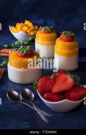 Traditional Italian sweet dessert Panna cotta with mango jelly served in jars and embellished with fresh real strawberrries and mint. Dark blue backgr Stock Photo