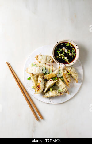 Fried asian dumplings Gyozas potstickers in white ceramic plate served with chopsticks and bowl of soy onion sauce over white marble background. Flat Stock Photo