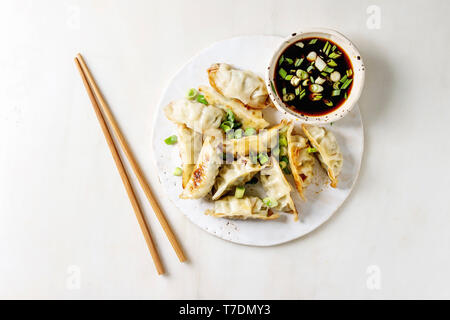 Fried asian dumplings Gyozas potstickers in white ceramic plate served with chopsticks and bowl of soy onion sauce over white marble background. Flat Stock Photo