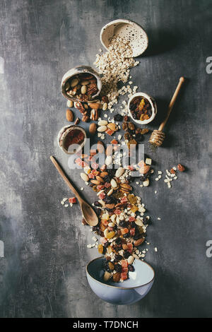 Variety of dried fruits, nuts, honey and oat flakes pours in ceramic bowl for cooking homemade healthy breakfast muesli or granola energy bars over bl Stock Photo