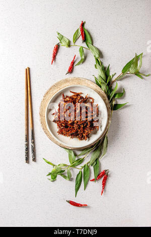 Vietnamese traditional hot spicy anchovies fish snack in ceramic plates with Vietnamese oregano greens and chopsticks over grey spotted background. Fl Stock Photo