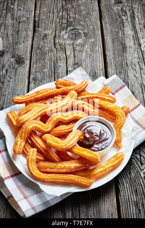 delicious churros with chocolate dipping  on a white plate on a wooden table with napkin, Traditional Spanish and Mexican street food, vertical view f Stock Photo