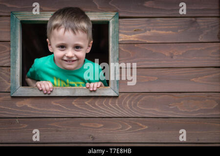 Portrait of a cute little Caucasian boy looking through the window of a wooden toy house in a outdoor playground Stock Photo