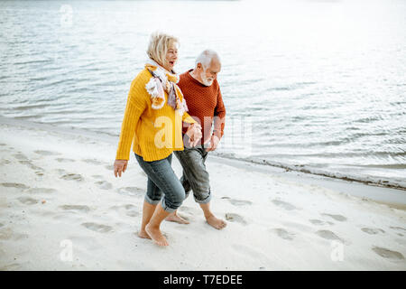Lovely senior couple dressed in colorful sweaters walking on the sandy beach, enjoying free time during retirement near the sea Stock Photo