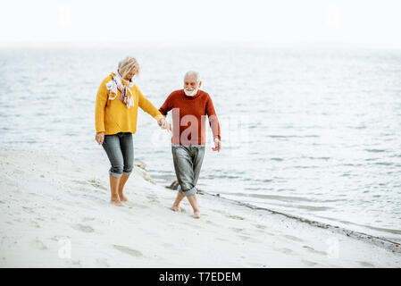 Lovely senior couple dressed in colorful sweaters walking on the sandy beach, enjoying free time during retirement near the sea Stock Photo