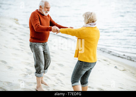 Lovely senior couple dressed in colorful sweaters dancing on the sandy beach, enjoying free time during retirement near the sea Stock Photo