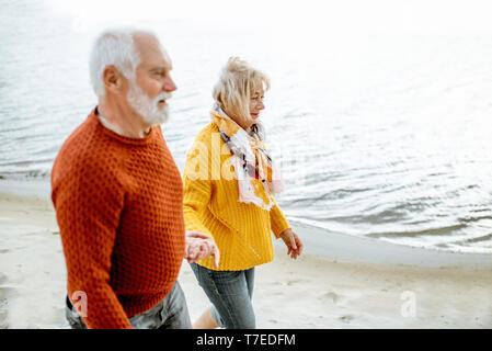 Lovely senior couple dressed in colorful sweaters walking on the sandy beach, enjoying free time during retirement near the sea Stock Photo