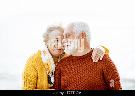 Portrait of a happy senior couple dressed in colorful sweaters hugging on the sandy beach, enjoying free time during retirement near the sea Stock Photo