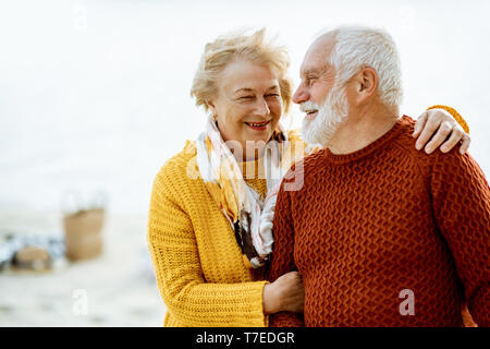 Portrait of a happy senior couple dressed in colorful sweaters hugging on the sandy beach, enjoying free time during retirement near the sea Stock Photo