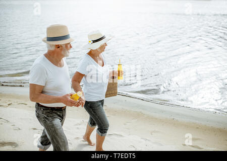 Happy senior couple dressed in white t-shirts and hats walking together on the sandy beach during their retirement Stock Photo