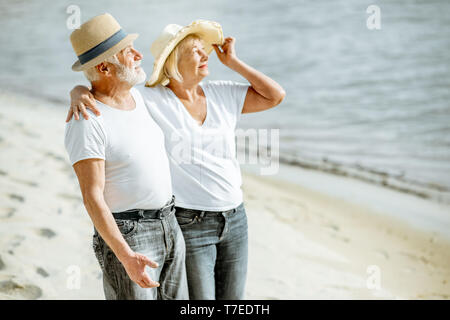 Happy senior couple dressed in white t-shirts and hats relaxing together on the sandy beach during their retirement Stock Photo