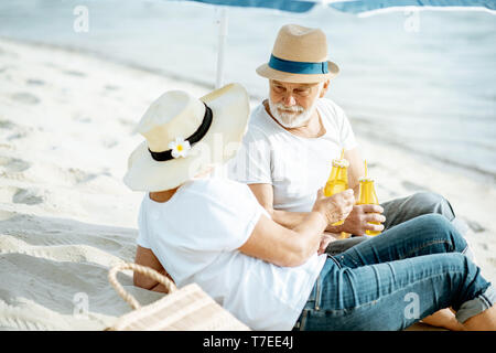 Happy senior couple relaxing, lying with drinks on the sandy beach, enjoying their retirement near the sea Stock Photo