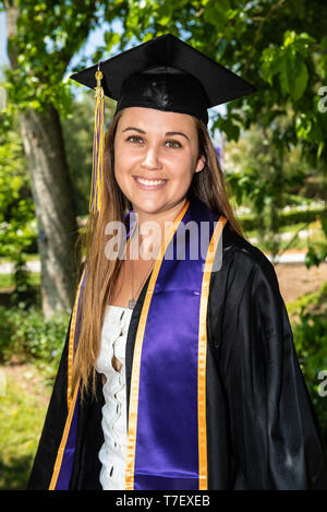 Beautiful young female student looking to left in close up view while wearing mortar board hat, tassel, and gown. Stock Photo