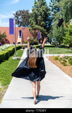 Beautiful young female student walking through campus in her gown while flying mortar board hat off to the side. Stock Photo
