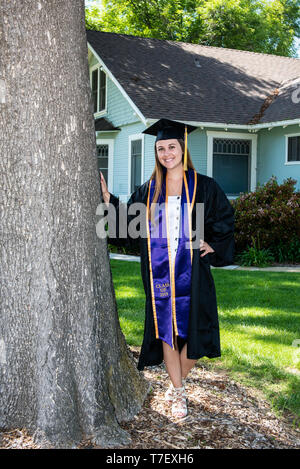 Beautiful young female student smiling with accomplishment while leaning against tree in front yard of home. Stock Photo