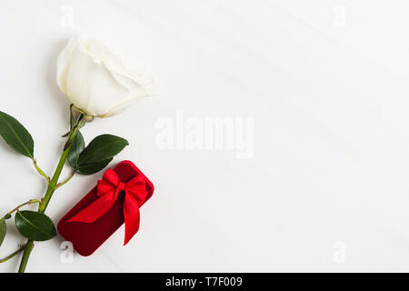 Red box for jewellery with red bow and white rose on white textured background. Valentine's Day or wedding concept. Sign of love. Copy space. Flat lay