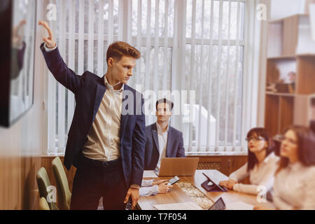 office workers like a team hold a meeting at one desk for laptops, tablets and papers, on the background a large TV set on a wooden wall Stock Photo
