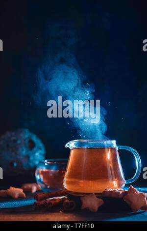 Glass teapot with rising steam and star-shaped cookies on a dark background with copy space. Astronomy themed Stock Photo