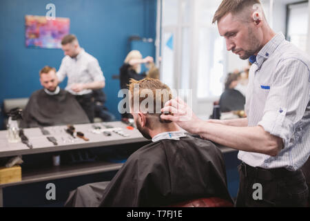 Close up shot of a man getting a trendy haircut at a hair salon. Male caucasian hairdresser serving a client. Man with a beard making haircut with a t Stock Photo