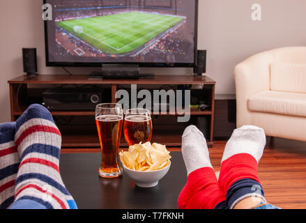 Relaxed man lying on sofa while watching football match on television at home, beer and chips on table Stock Photo