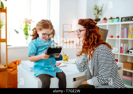 Loving red-haired mom looking at funny daughter using calculator Stock Photo