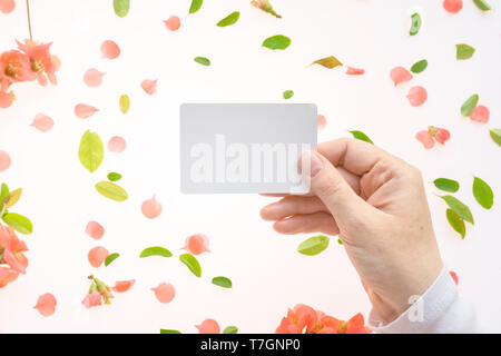 Woman holding blank white business card mock up in hand over white background with blooming springtime flower petals and leaves Stock Photo